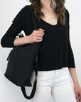 Close up view of a brown haired model facing the camera. She is wearing a black shirt and white pants. She carries Sherpani's Anti-Theft bag, the Vale AT in Carbon, over her shoulder as a tote.