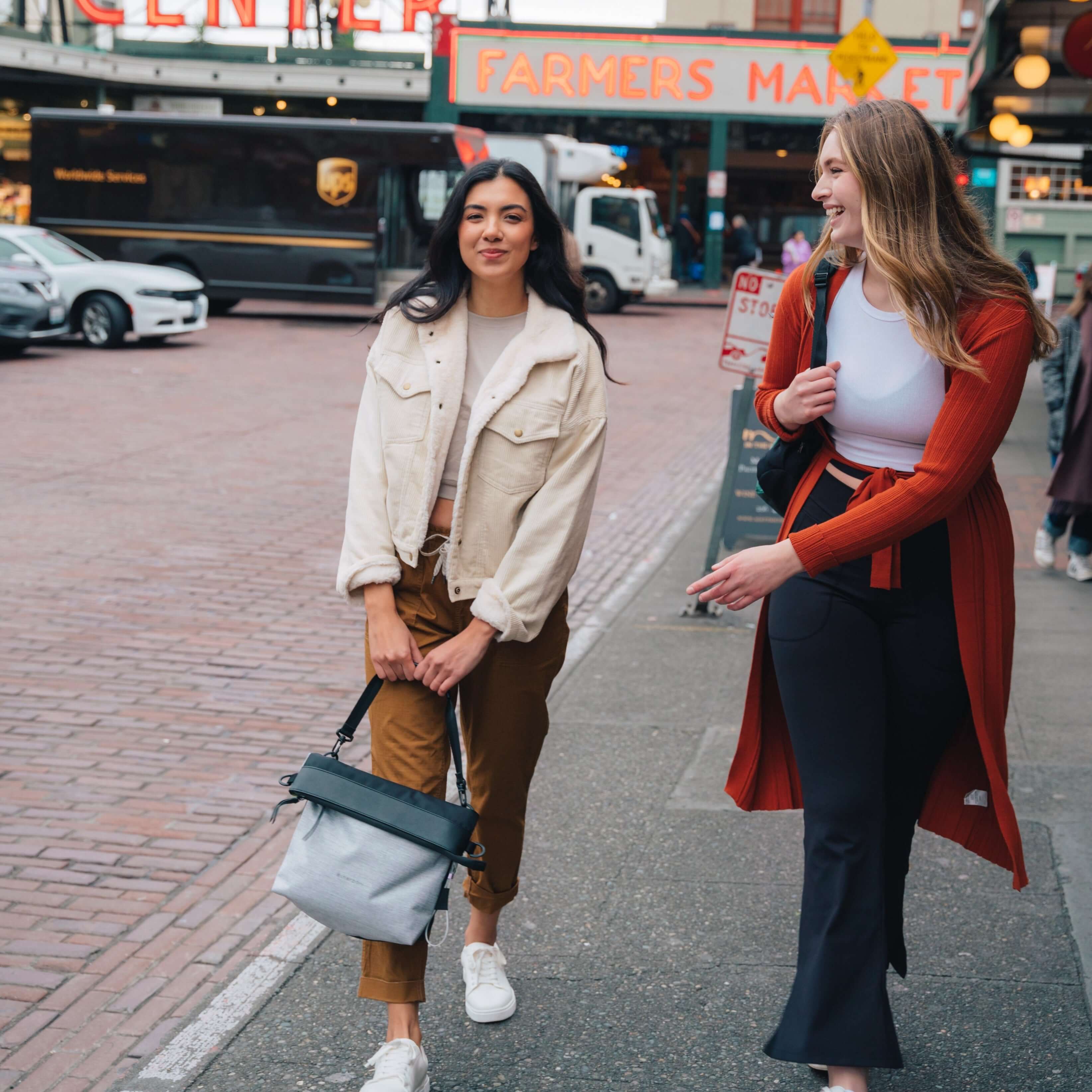 Two women are walking outside in a market and smiling. The dark haired woman on the left is wearing a white jacket and orange pants. She is grabbing the handles of Sherpani&#39;s Anti-Theft tote, the Vale AT in Sterling. 