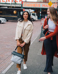 Two women are walking outside in a market and smiling. The dark haired woman on the left is wearing a white jacket and orange pants. She is grabbing the handles of Sherpani's Anti-Theft tote, the Vale AT in Sterling. 