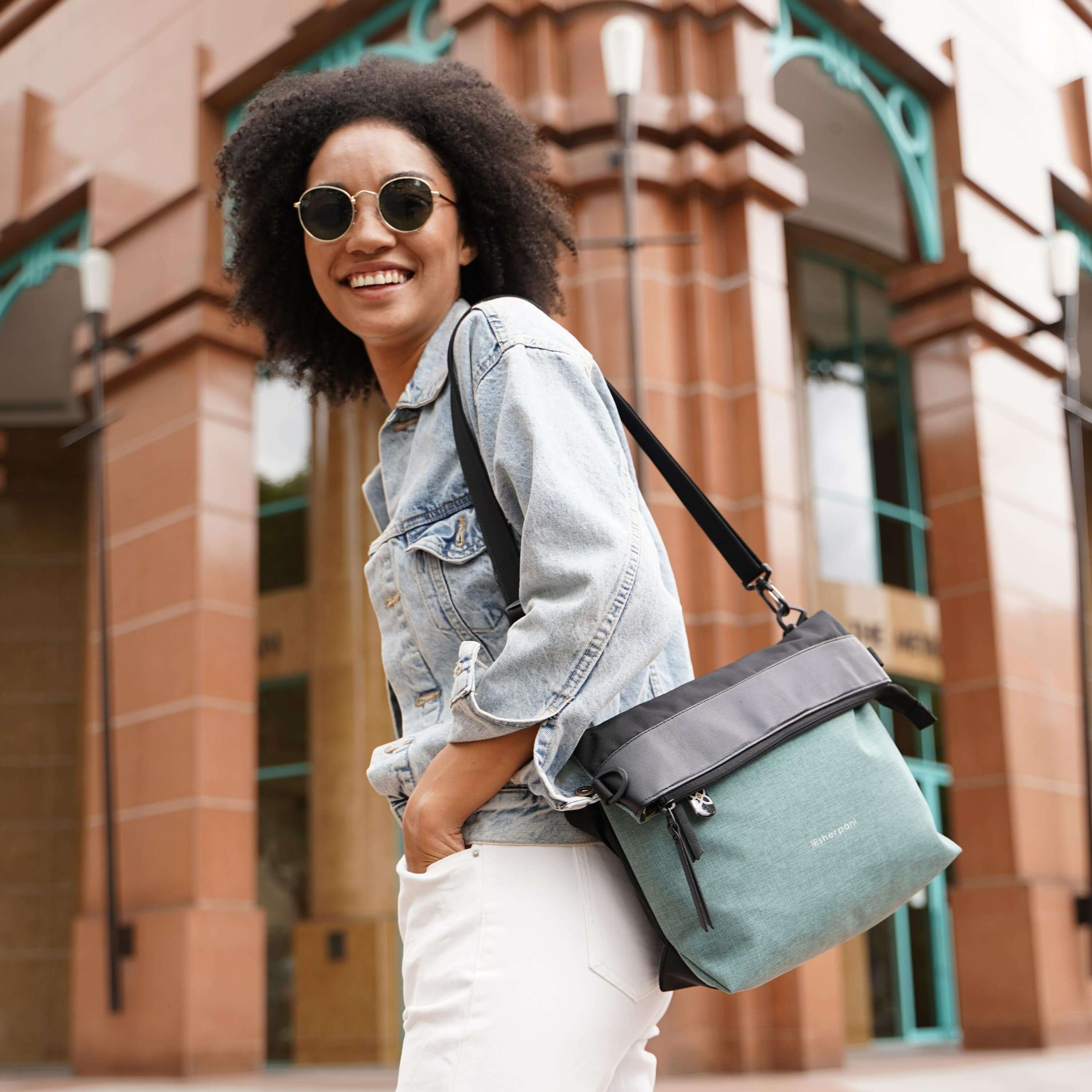 Close up view of a curly haired model looking over her left shoulder and smiling at the camera. She is wearing sunglasses, a jean jacket, white pants and Sherpani's Anti-Theft bag, the Vale AT in Teal, over her shoulder.