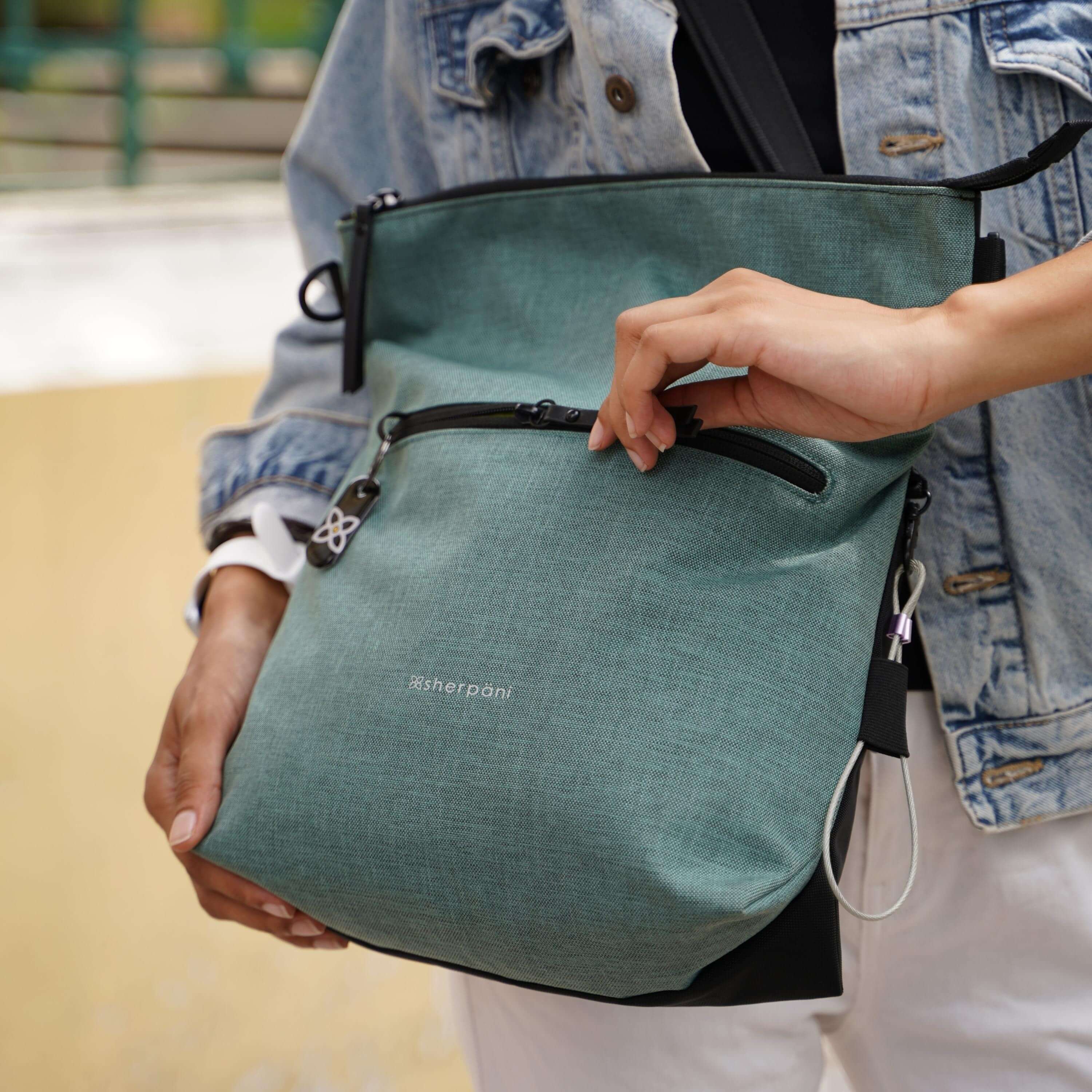 Close up view of a model&#39;s hands. She is unzipping the external compartment of Sherpani&#39;s Anti-Theft bag the Vale AT in Teal.