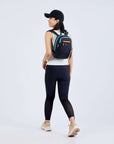 Full body view of a dark haired model facing away from the camera and smiling over her left shoulder. She is wearing a black ball cap, white tank top, black leggings and white sneakers. She carries Sherpani mini backpack, the Vespa in Chromatic.