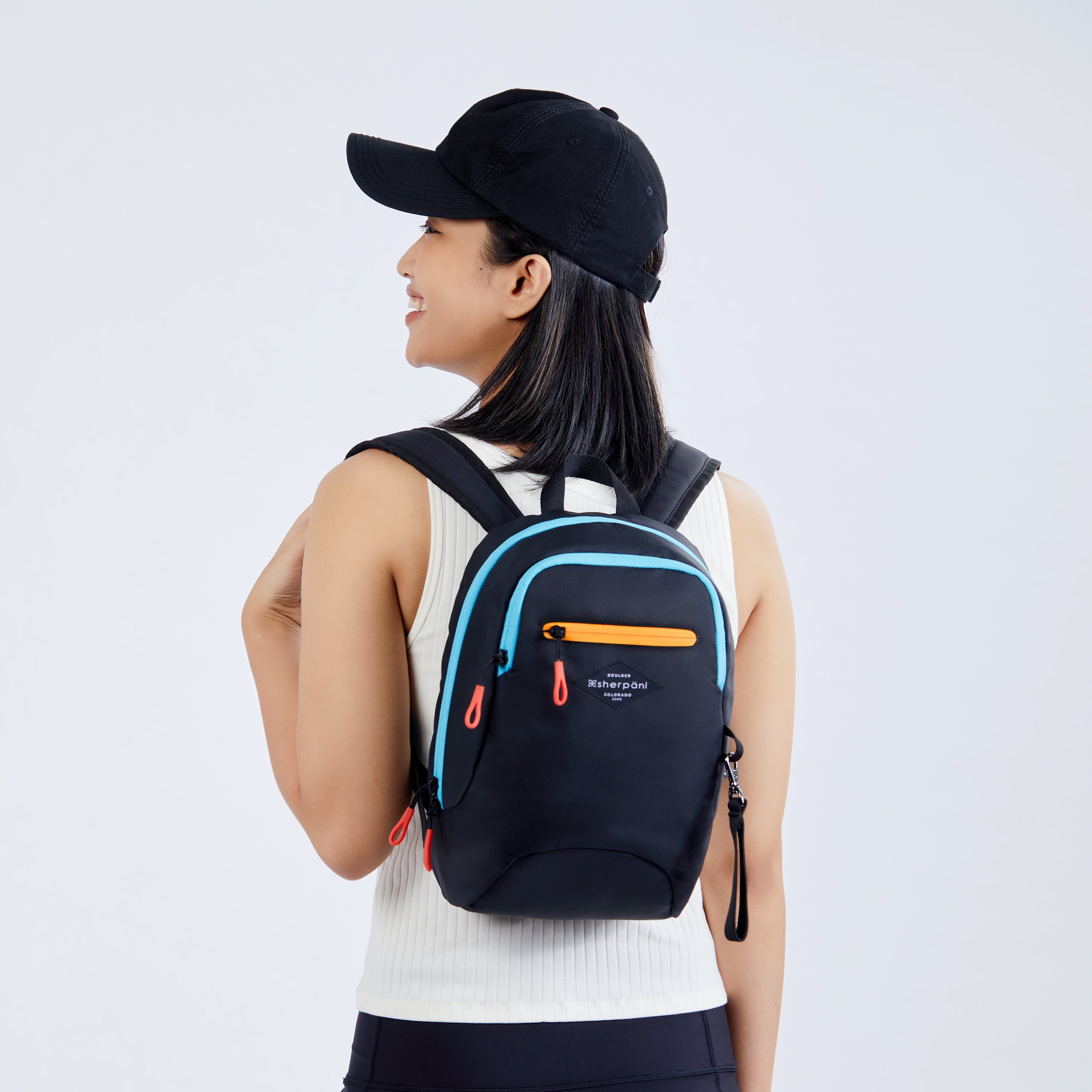 Close up view of a dark haired model facing away from camera and smiling over her left shoulder. She is wearing a black ball cap, white tank top and black leggings. She carries Sherpani mini backpack, the Vespa in Chromatic.