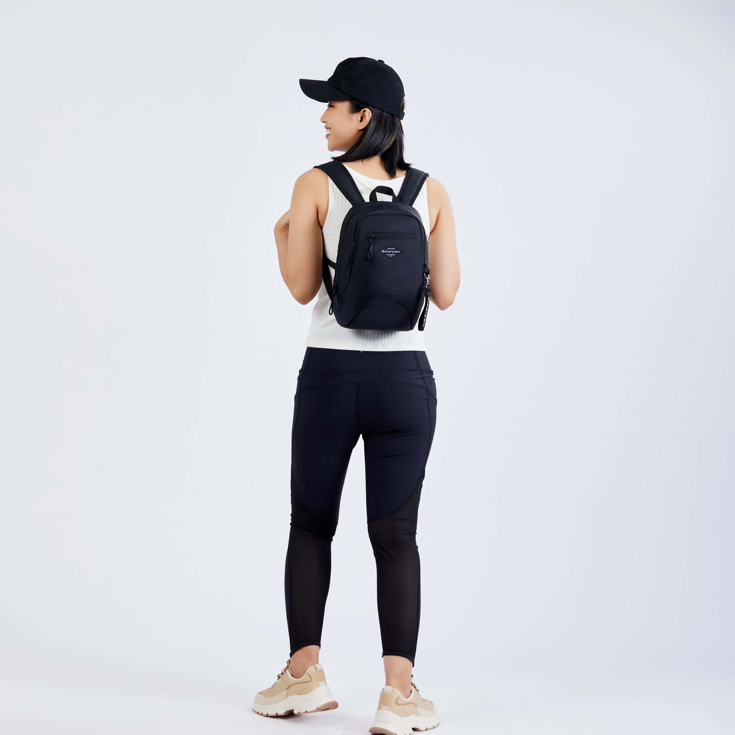 Full body view of a dark haired model facing away from the camera and smiling over her left shoulder. She is wearing a black ball cap, white tank top, black leggings and white sneakers. She carries Sherpani mini backpack, the Vespa in Raven.