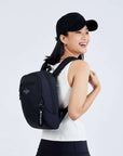 Close up view of a dark haired model facing away from camera and smiling over her right shoulder. She is wearing a black ball cap, white tank top and black leggings. She carries Sherpani mini backpack, the Vespa in Raven.