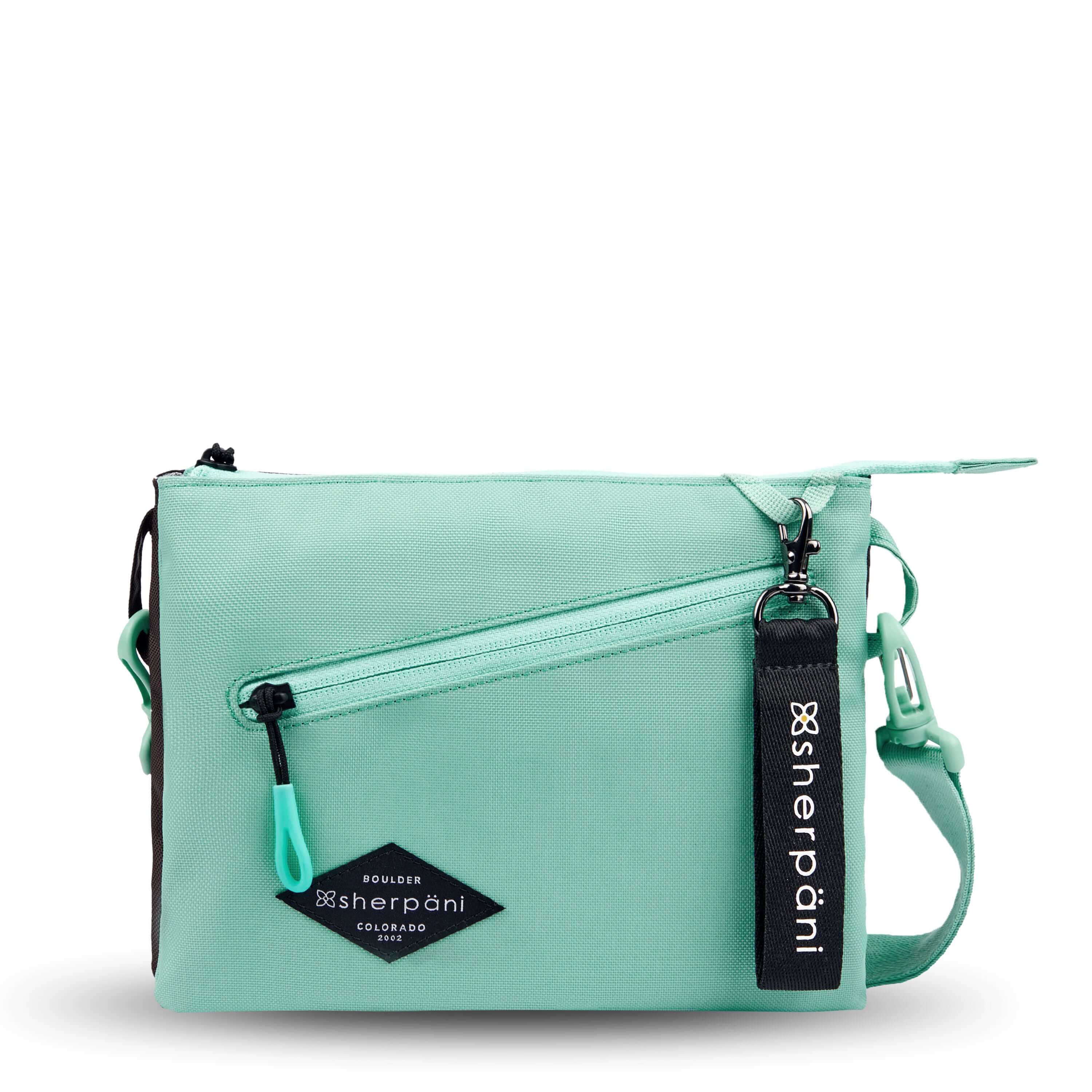 Flat front view of Sherpani crossbody, the Zoom in Seagreen. The bag is two-toned; the front half is light gray and the back half is brown. There is an external zipper pocket on the front with an easy-pull zipper accented in light green. The bag has an adjustable/detachable crossbody strap. There is a branded Sherpani keychain clipped to a fabric loop on the upper right corner. 