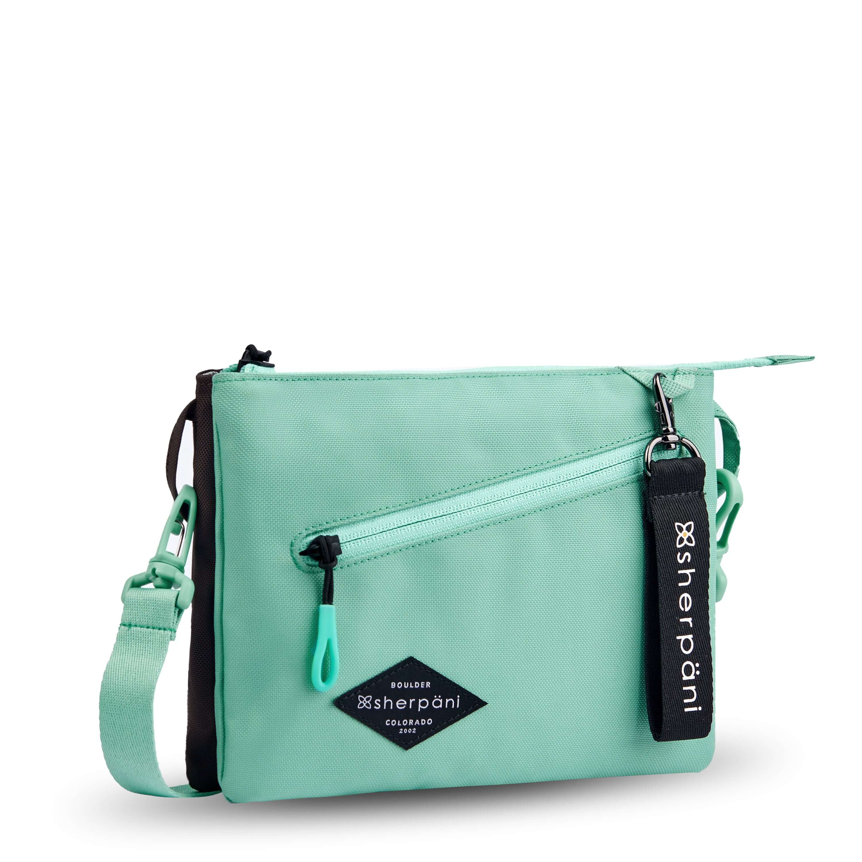 Angled front view of Sherpani crossbody, the Zoom in Seagreen. The bag is two-toned; the front half is light gray and the back half is brown. There is an external zipper pocket on the front with an easy-pull zipper accented in light green. The bag has an adjustable/detachable crossbody strap. There is a branded Sherpani keychain clipped to a fabric loop on the upper right corner. #color_seagreen