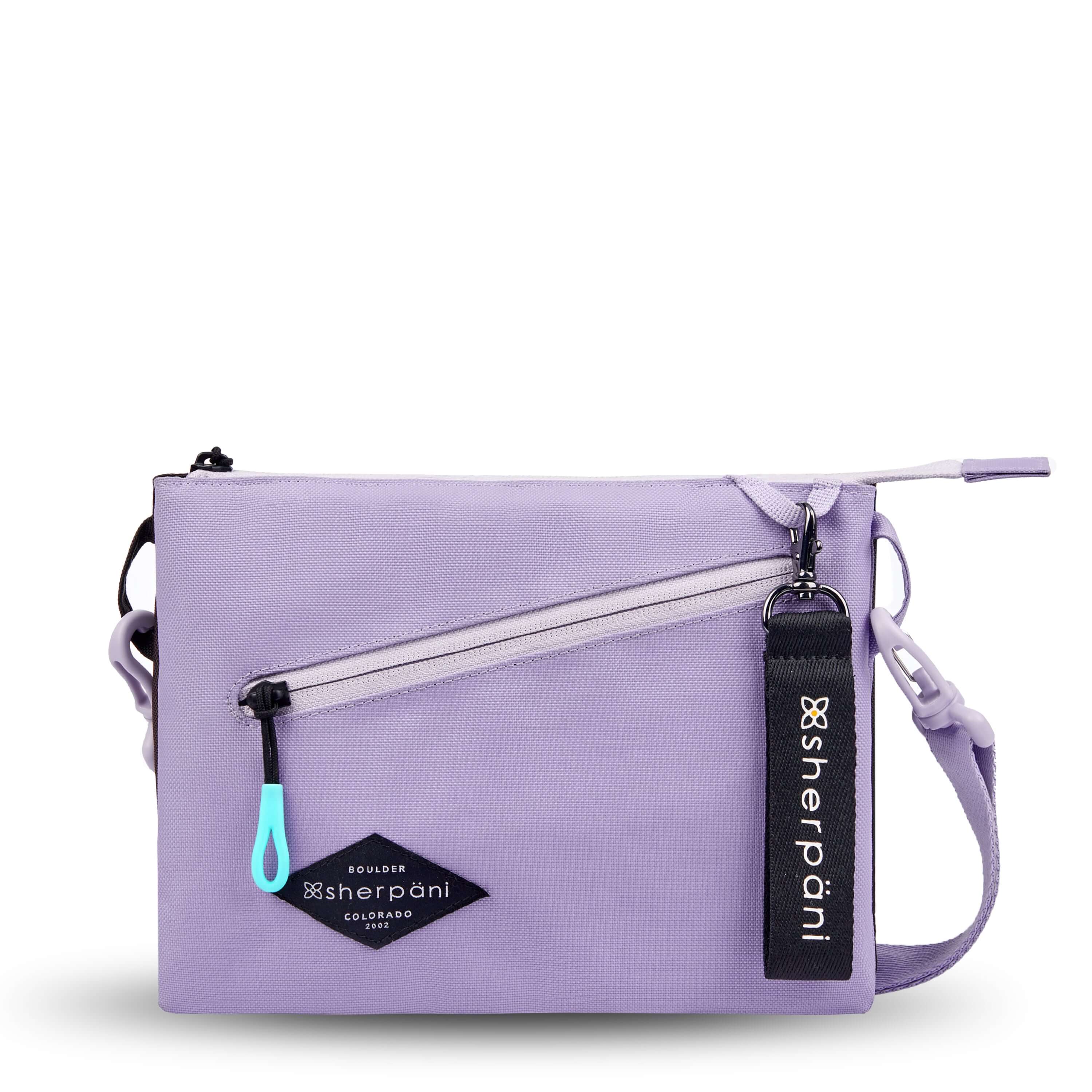 Flat front view of Sherpani crossbody, the Zoom in Lavender. The bag is two-toned; the front half is lavender and the back half is brown. There is an external zipper pocket on the front with an easy-pull zipper accented in aqua. The bag has an adjustable/detachable crossbody strap. There is a branded Sherpani keychain clipped to a fabric loop on the upper right corner. 