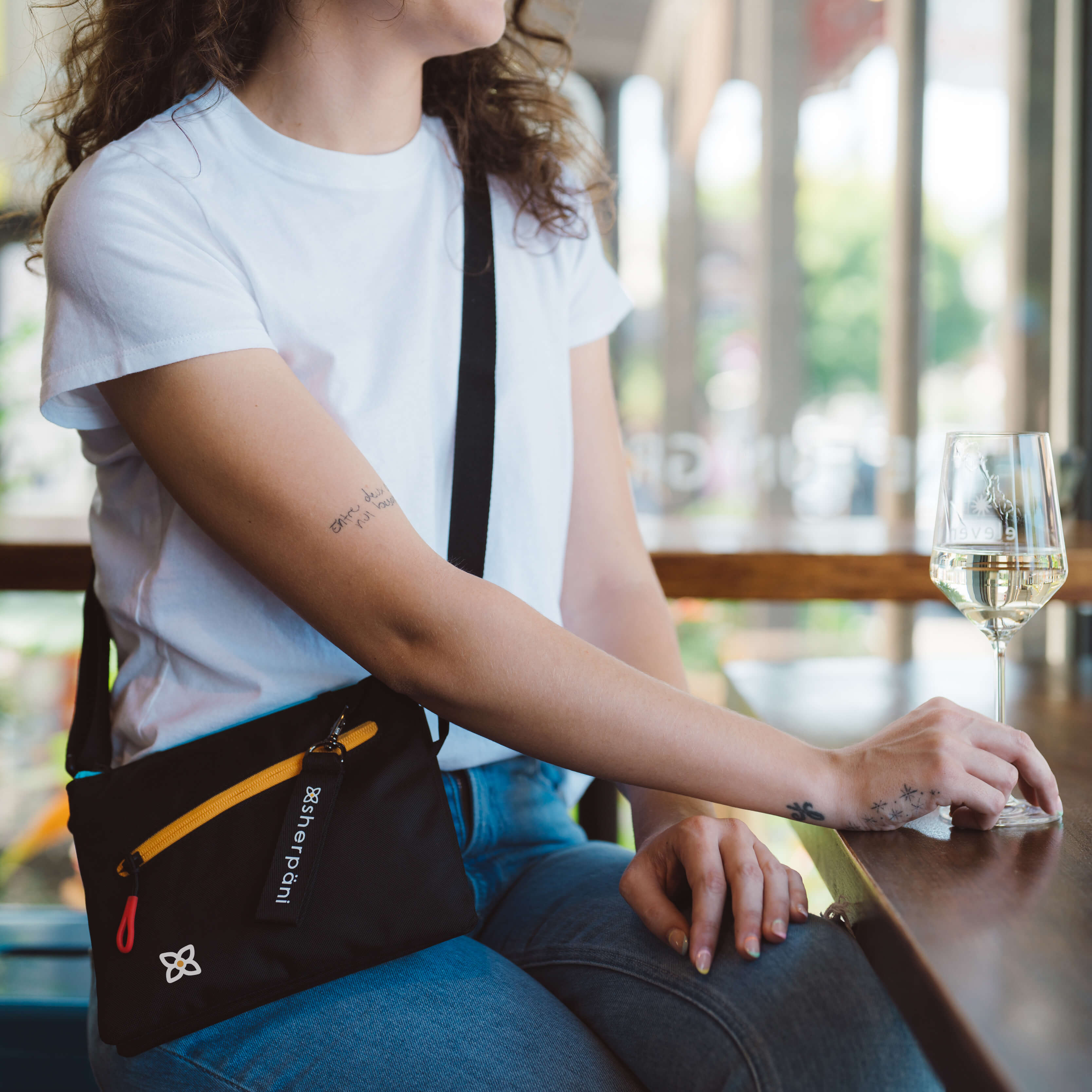 A woman sitting at a table drinking a glass of wine. She is wearing Sherpani RFID blocking bag, the Zoom in Chromatic. 