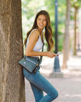 A woman outside leans against a tree. She is wearing Sherpani RFID blocking travel purse, the Zoom in Juniper.