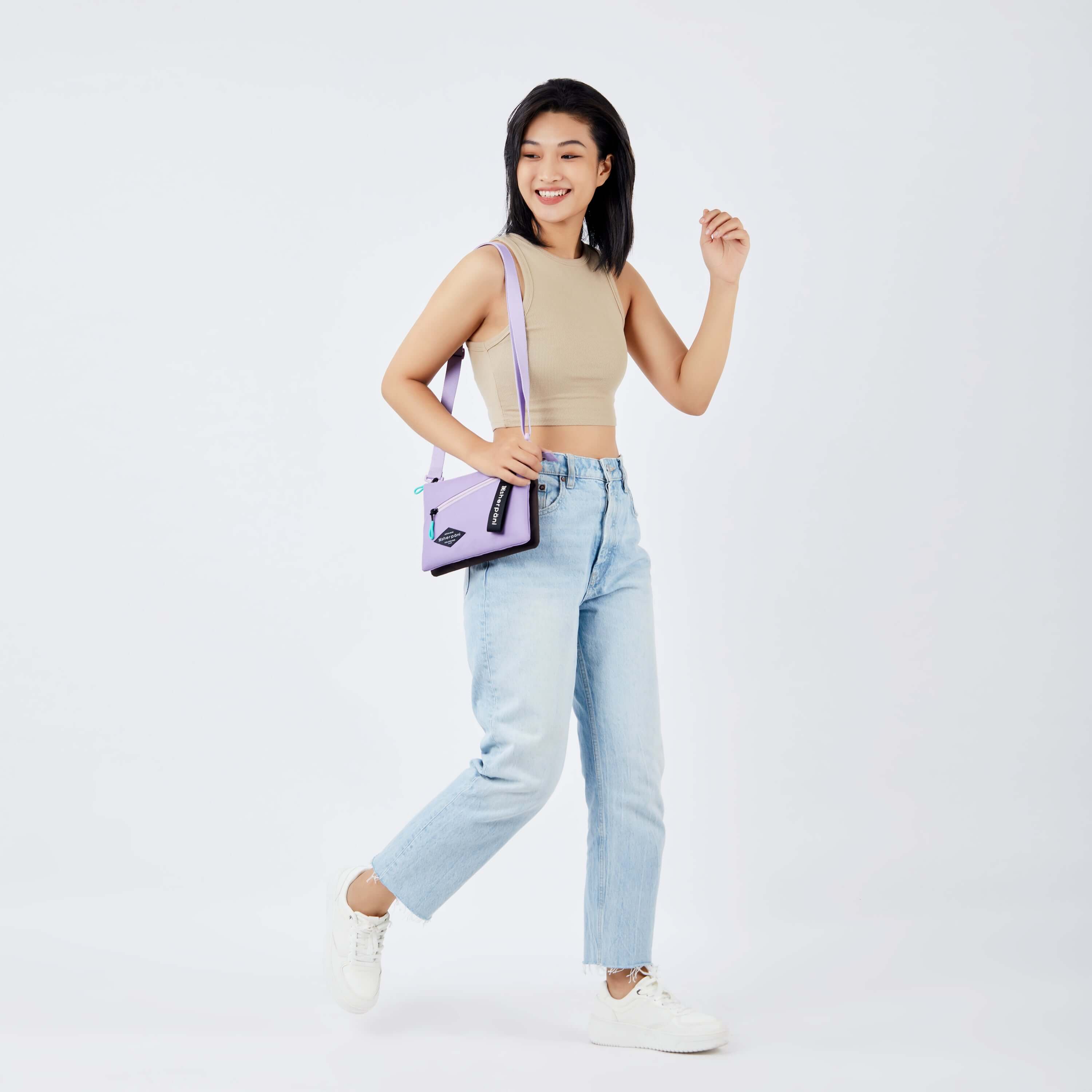 Full body view of a dark haired model stepping to the side and smiling. She is wearing a beige crop top, jeans and white sneakers. She carries Sherpani&#39;s crossbody, the Zoom in Lavender, over one shoulder.
