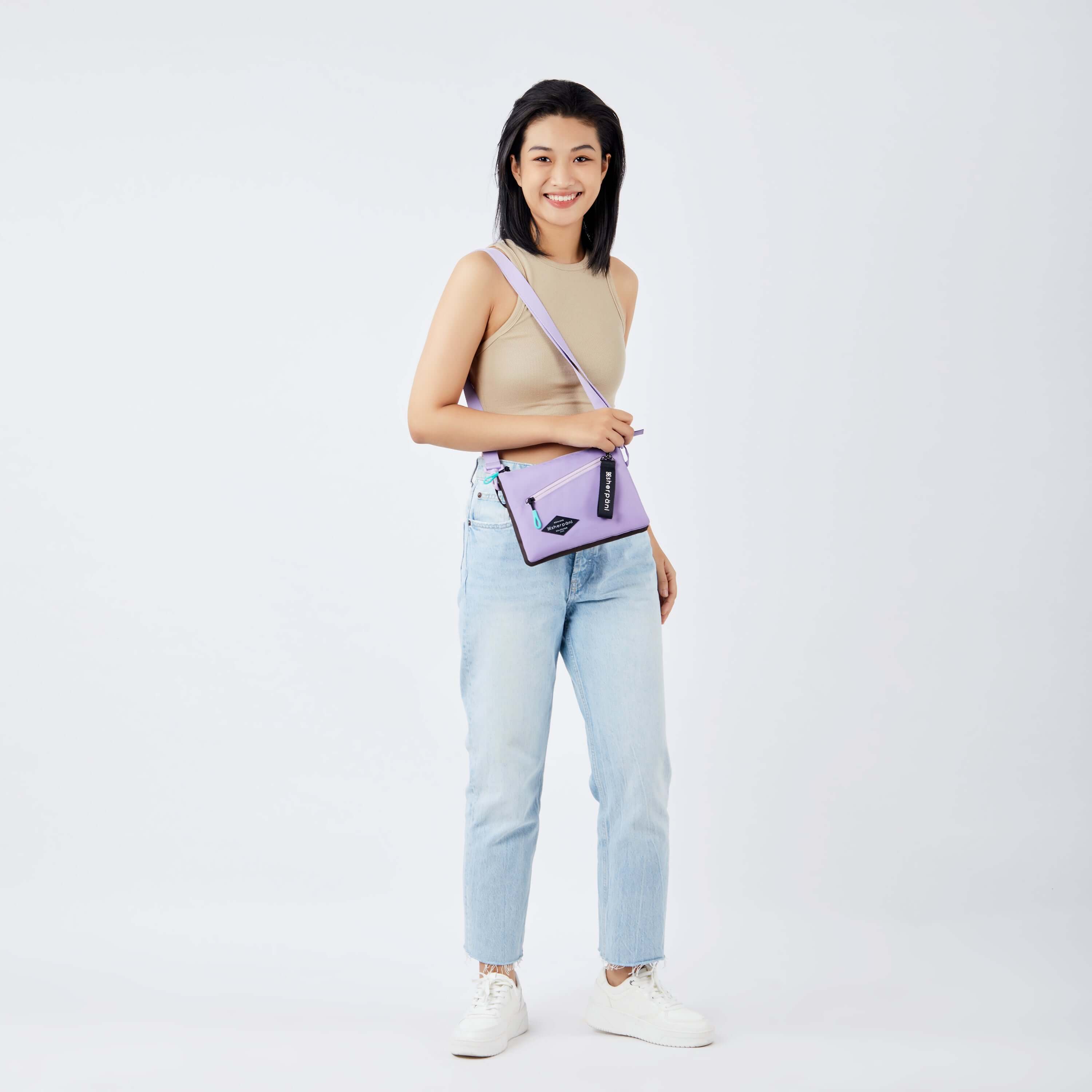Full body view of a dark haired model facing the camera and smiling. She is wearing a beige crop top, jeans and white sneakers. She carries Sherpani&#39;s crossbody, the Zoom in Lavender, over one shoulder.