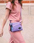 Close up view of a red haired woman walking outside in front of a brick wall. She is wearing a salmon pink top and matching pants. She carries Sherpani crossbody, the Zoom in Lavender.