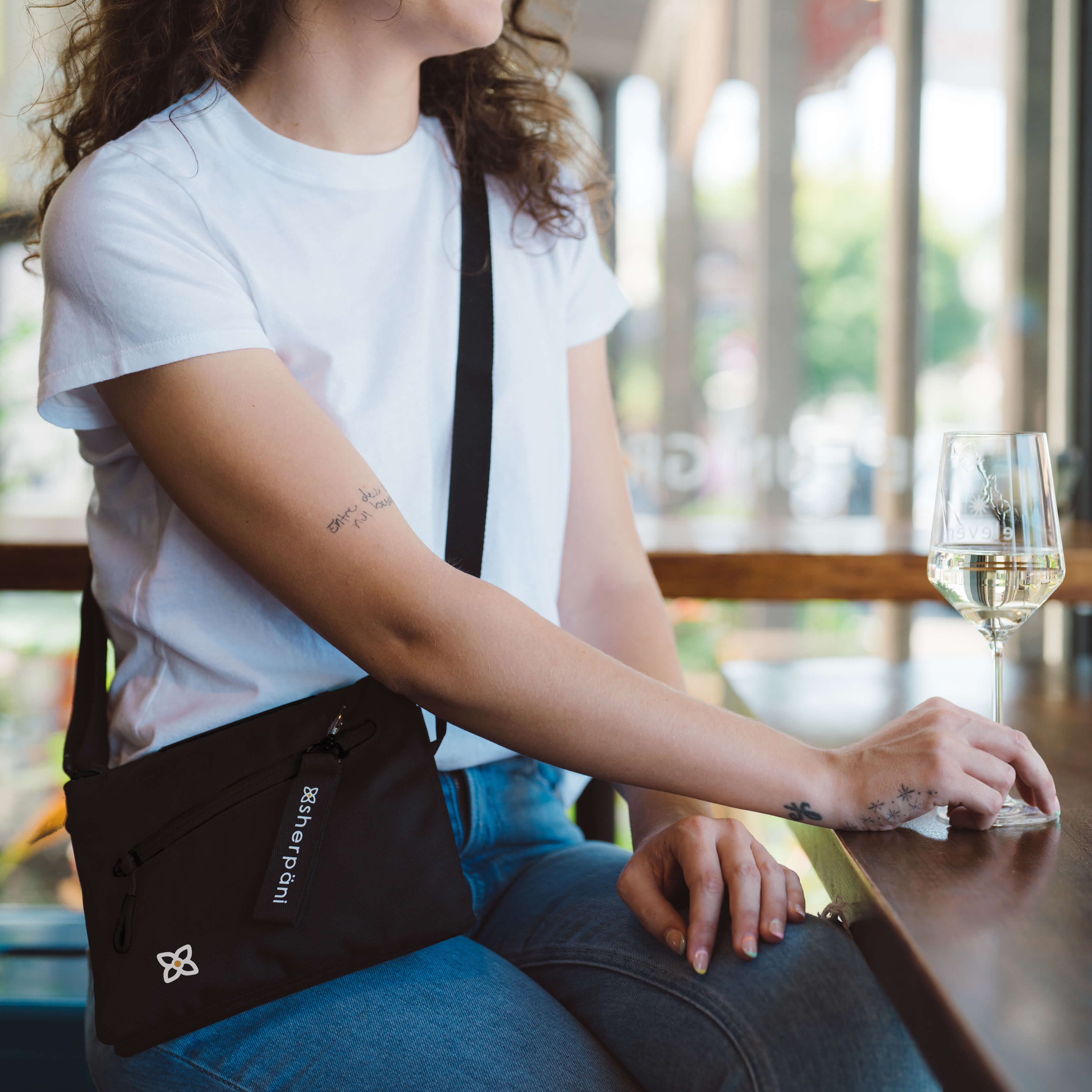 A woman sits at a table with a glass of wine. She is wearing Sherpani RFID blocking bag, the Zoom in Raven.