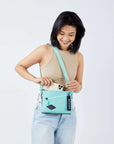 Close up view of a dark haired model facing the camera and smiling downward. She is wearing a beige crop top and jeans. She is pulling her phone of out of Sherpani crossbody, the Zoom in Seagreen, which is over her shoulder.