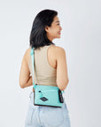 Close up view of a dark haired model facing away from the camera and smiling over her right shoulder. She is wearing a beige crop top and jeans. She carries Sherpani crossbody, the Zoom in Seagreen, as a crossbody.