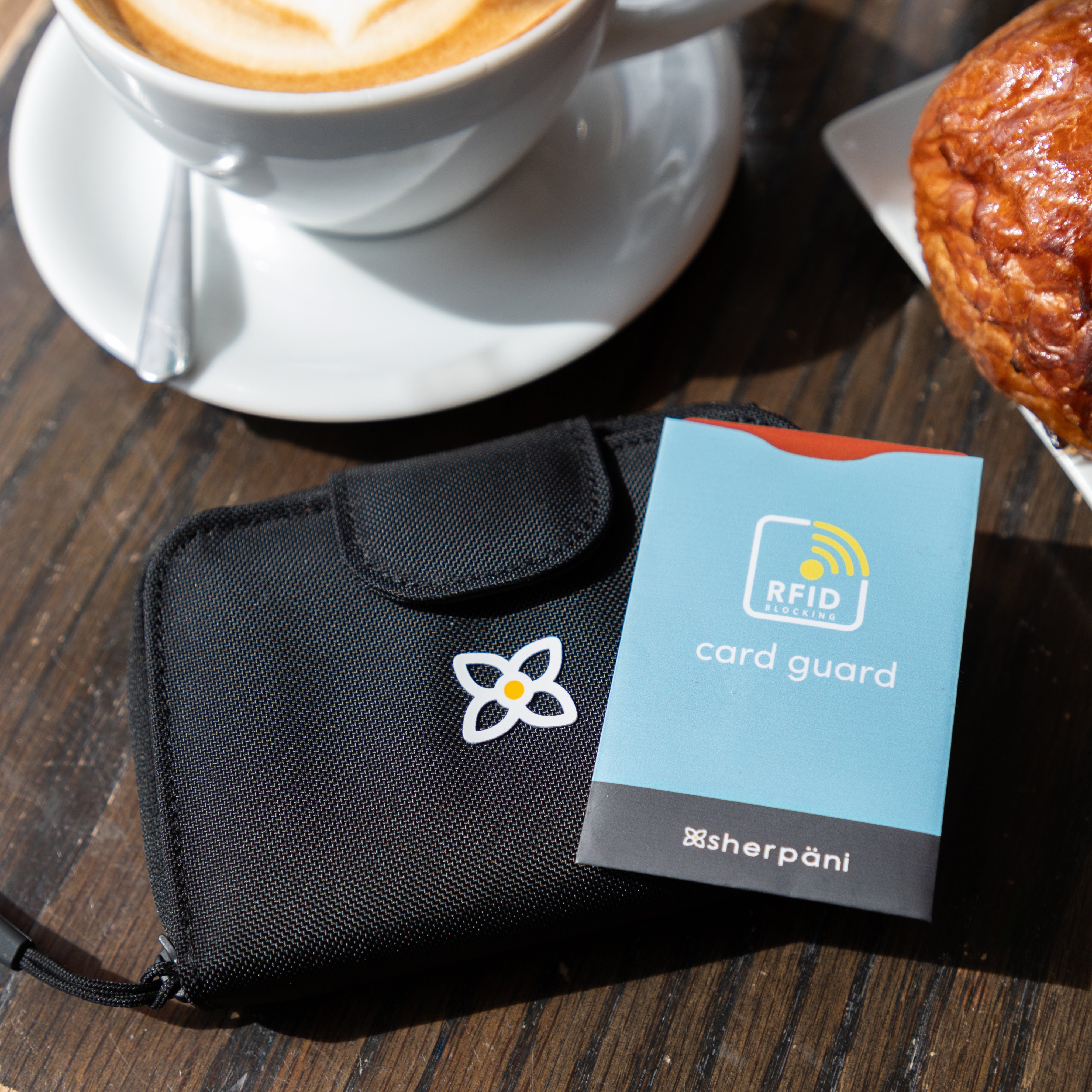 On top of a table with a coffee and a pastry, the card guard in Maui Blue sits on top of Sherpani RFID wallet, the Barcelona.