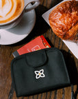 On top of a table with a coffee and a pastry, the card guard in Cider peeks out from Sherpani RFID wallet, the Barcelona.